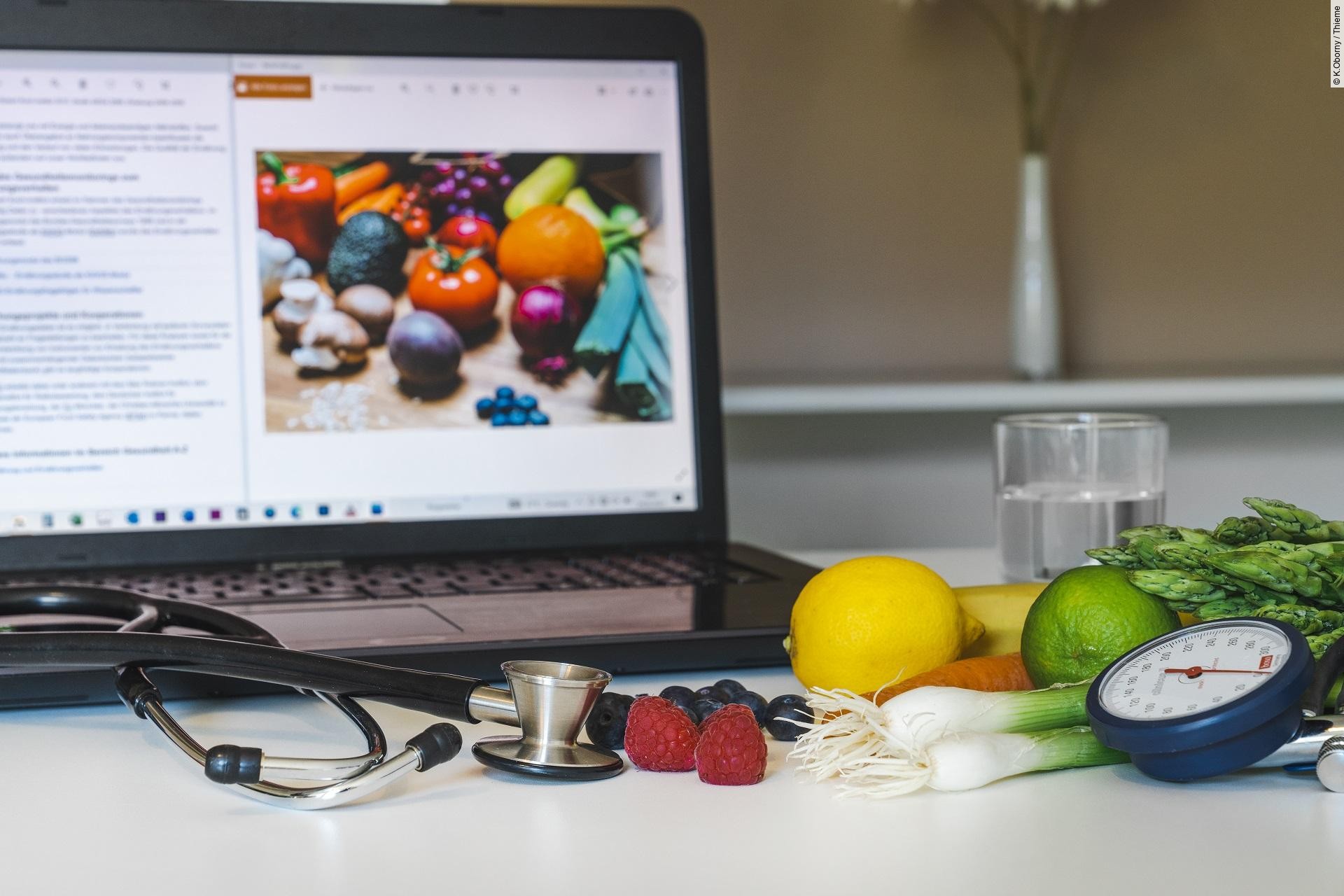 Laptop, der umgeben ist von Obst, Gemüse, einem Stethoskop und Blutdruckmessgerät.