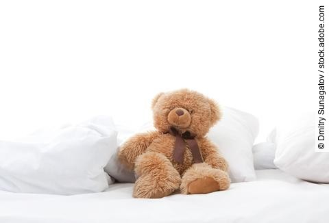 Teddybär sitzt auf einem Bett mit weißen Laken.