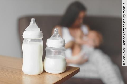 Zwei Flaschen mit Muttermilch stehen auf einem Tisch. Im Hintergrund hält eine Mutter ihr Baby. 