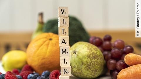 Obst und Gemüse im Hintergrund und Buchstaben auf Würfeln, die aufeinandergestapelt das Wort "Vitamine" ergeben.